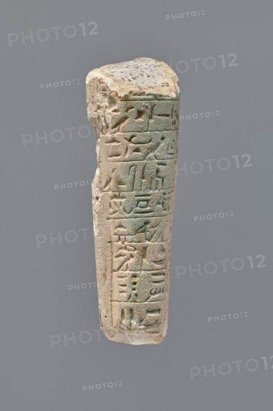 Ushabti - fragment, unknown role classification of creator, Mummy-shaped figurine, grave good. Fragment of the legs of an ushabti preserved from hips to ankles. The back rests on a pillar. Inscribed hieroglyphs placed in ten horizontal bands around the legs, separated by thin lines. 332–30 BC; Ptolemaic Period. Tell Atrib; ushabti. Height 8.5 cm, width 3.8 cm., A weathered vertical stone fragment with carved grooves and shallow reliefs, showing a worn top edge and an irregular side profile; the surface displays a mix of light beige, sandy tan, pale gray, and areas of greenish patina, with subtle darker brown spots and shadows accentuating the carved lines and recessed areas.