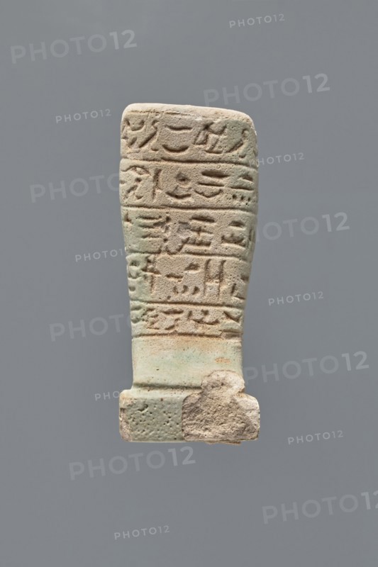 Ushabti - fragment, Mummy-shaped figurine, grave good. Fragment of the legs preserved from the knees down; on the back a pilaster, the feet on a base. Imprinted hieroglyphs in five bands around the legs. 332–30 BC; Ptolemaic Period. Found at Tell Atrib. Ushabti. Height 7.2 cm, width 3 cm., A vertical carved object with a rectangular top tapering slightly toward a wider base, showing a weathered surface with shallow incised marks arranged in horizontal bands; the material appears grainy and worn with areas of rougher texture and small pits, and a chunk missing near the lower right edge; the dominant colors visible are pale beige, light tan, and soft green, set against a plain neutral gray background.