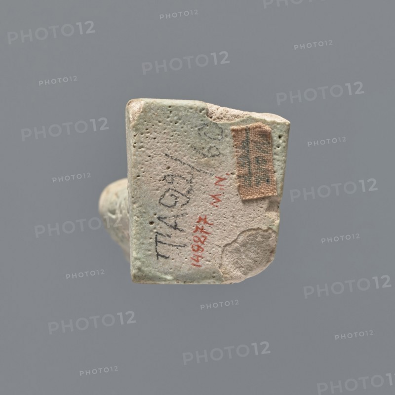 Ushabti fragment, mummy-shaped figurine; fragment of the legs preserved from the knees down. On the back there is a pilaster; the feet on a base. Imprinted hieroglyphs in five bands around the legs. Ushabti used as grave goods. Ptolemaic Period, 332–30 BC. Tell Atrib. Materials/dimensions: height 7.2 cm, width 3 cm., A small worn rectangular object with rounded edges is shown against a gray background; its surface is textured with small pits and irregularities and displays a worn pale greenish-beige color with areas of light beige and off-white; a small rough patch of tan fabric is attached to one corner, and there are smudges and darker gray marks on the surface; the object appears chipped at one edge, revealing a lighter interior, and a faint shadow is cast beneath it.