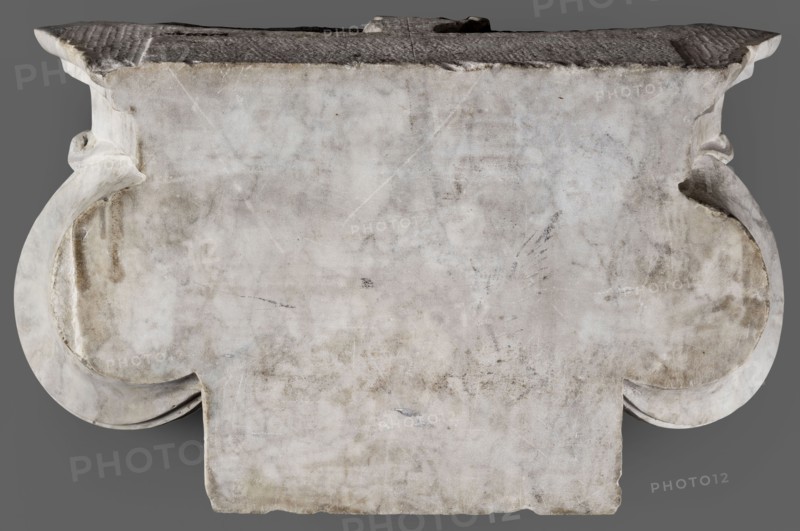 Ionic pilaster capital, unknown Roman workshop, 2nd century, Roman Empire 27 BC–476, architectural element; materials/dimensions: height 27 cm also listed 26 cm, width 46.5 cm, depth 18 cm, weight 19.5 kg., A weathered stone slab with a broad, rectangular top and ornate curved sides sits against a neutral gray background, the stone surface showing a mottled pattern of off-white, gray, and beige with darker smudges and faint scratches; the top edge appears darker and rougher with textured ridges, and the curved side elements flare outward with smoother, slightly shadowed undersides, while the front face displays subtle discoloration, worn patches, and irregular edges along the bottom.