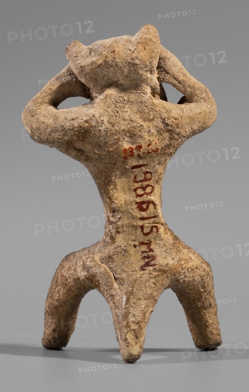 Satyr figurine, clay; depicts a satyr with a face stylized as a mask with protruding ears and two horns. The crouching male figure has legs widely apart, supported by a thick tail; hands are raised and touch the head tilted slightly left, emphasizing and exposing the genitals. Face with lush, regular facial hair; small eyes, clearly defined eyebrows and broad nose, thin closed mouth. Hands and legs are treated schematically compared to the carefully modeled face. Made of fine-grained yellowish-tinted clay. Date: 5th century BC previous datings: 2nd half of the 6th century BC–beginning of the 5th century BC; earlier: end of the 6th–first half of the 5th century BC. Place of origin: Corinth. Figurine; height 8.8 cm, width 4.8 cm, depth 4.4 cm; weight 0.04 kg., A small rough-textured figurine with a weathered surface in shades of beige, tan, and brown, shown from the back; the figure has a broad upper body with two raised arms bent so hands rest near the head, a narrow waist, and a lower ...
