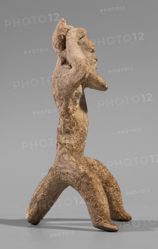 Satyr figurine. Clay figurine depicting a satyr with a face stylized as a mask with protruding ears and two horns. The figure crouches with legs widely apart, supported by a thick tail; hands are raised upwards, touching the head tilted slightly to the left and emphasizing the exposed genitals. Face has lush, regular facial hair; small eyes; clearly defined eyebrows and broad nose; thin closed mouth. Hands and legs are treated schematically compared with the carefully modeled face. Fine-grained yellowish clay. 5th century BC previously dated to late 6th–early 5th century BC. Origin: Corinth. Medium: clay figurine. Dimensions: height 8.8 cm; width 4.8 cm; depth 4.4 cm; weight 0.04 kg., A weathered tan and brown clay figure standing on a flat gray surface against a smooth gray background, with a rough textured surface and uneven coloration; the figure has a vertical torso, elongated bent legs forming a wide stance, and raised arms positioned near its head, with a protruding facial profil...