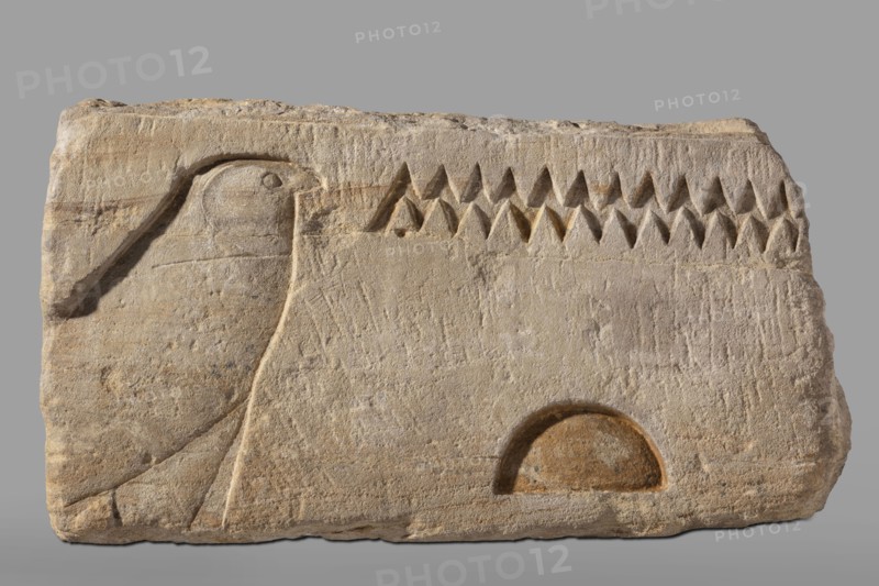 Fragment of an architrave with hieroglyphs from the temple of Horus in Buhen. Horus mythology, falcon iconography, hieroglyphs, Egyptian sacred architecture. Fragment of an architrave with preserved hieroglyphs in recessed relief. From the left, the sign hor, followed by the signs n and t. Traces of yellow polychrome are preserved on the surface. c. 1479–c. 1425 BC; New Kingdom, 18th Dynasty possibly Thutmose III. Findspot: Buhen, Faras site/country of find. Bas-relief, architectural element. Height 31 cm; width 60 cm; depth 52 cm; weight 143.5 kg., A rectangular stone slab in beige and light brown tones with rough, weathered texture shows a low-relief carving of a stylized bird profile on the left, with a rounded head, beak, and a simple carved eye, and a gently curved body and wing lines; across the upper center is a row of carved triangular shapes creating a sawtooth pattern in darker brown shadows; near the lower right there is a semicircular recessed area with warmer brown shading...