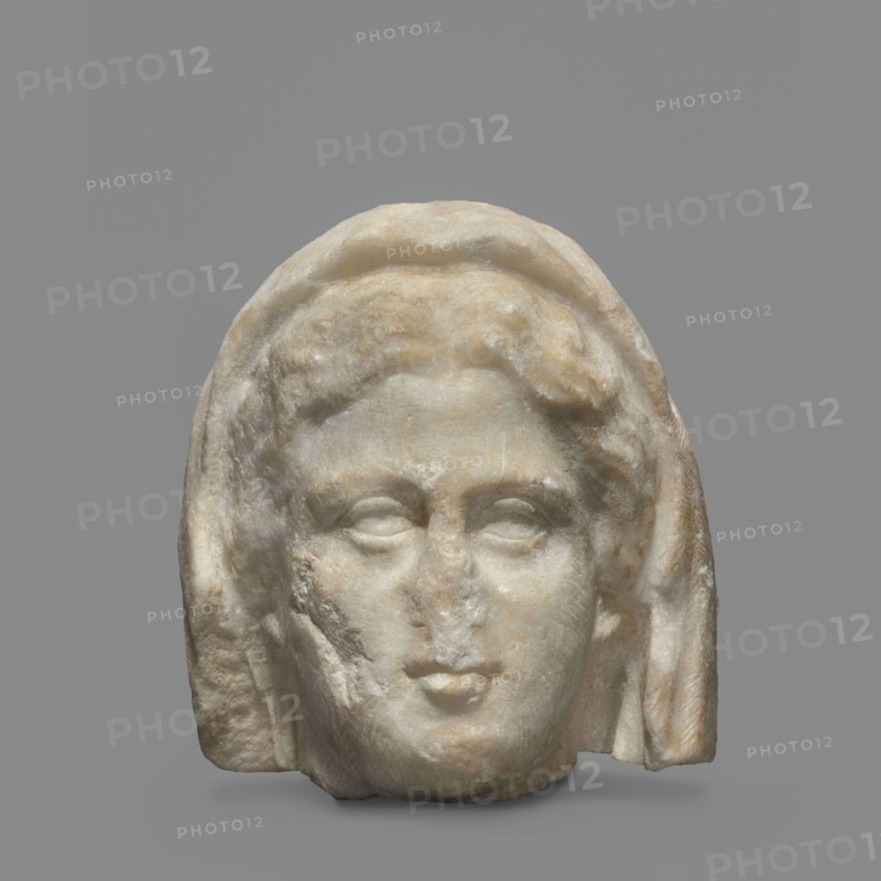 Head of a woman wearing a veil. The face is oval with regular features: rounded chin, small compact mouth, and slim cheeks. From the nose, skin folds extend diagonally downward across the cheeks. The eyes are large, open, deeply set, with thick upper and lower eyelids; outer and inner corners are level. Eyeballs are convex with dimpled pupils. Eyebrows are cylindrical, sloping toward the temples. Forehead is high, convex above the nose and slightly sunken above the eyebrows. Hair is lush, wavy, parted above the forehead and combed over the temples, revealing the lower parts of the ears. The head is covered with a veil. Head in semi-profile, 1st half of the 4th century BC, Athens; bas-relief, height 12 cm, width 11 cm, depth 11 cm., A carved stone head of a person with a rounded forehead and stylized hair partially covered by a draped hood, rendered in worn pale beige and off-white stone with patches of light tan and subtle gray veining; the face shows softly modeled features including ...