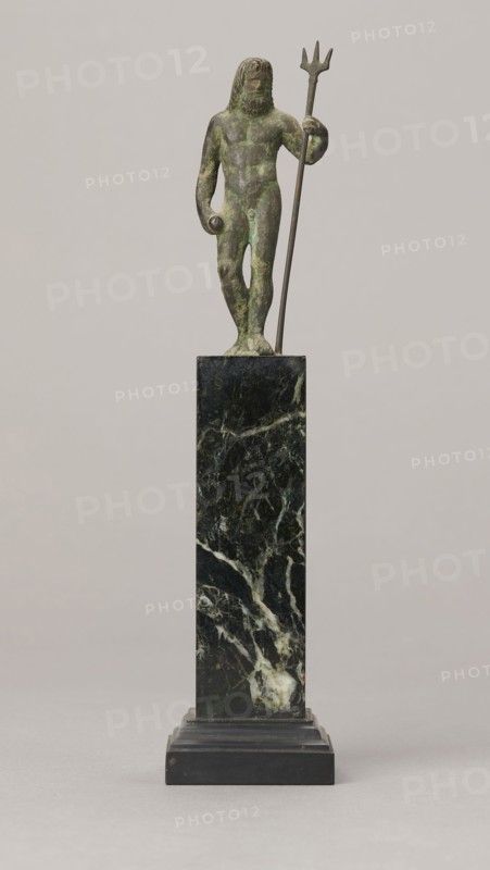 Figurine of Poseidon, unknown Roman workshop. Figurine of Poseidon standing in contrapposto, resting on a trident. The weight of the body rests on the left leg; the right is slightly moved back, the foot bent at the toes. The body is nude, correctly modeled, with clearly defined abdominal muscles. In the left hand the figure holds a trident; the right hand is lowered at the side of the body, holding a cylindrical object at hip level. The head is slightly bowed. The face is framed by long hair and a beard. 1st century, Roman Empire 27 BC–476. Bronze figurine; height 8.7 cm, width 3.5 cm, depth 1.8 cm, weight 0.362 kg with base., A small greenish bronze figure with a patina stands on a tall dark green and black marble pedestal; the figure holds a long thin trident in one hand and a small rounded object in the other, has long hair and a beard, and shows muscular body contours in shades of green and brown; the pedestal displays veining in white and lighter green against a dark background a...