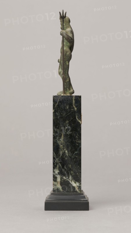 Figurine of Poseidon, unknown Roman workshop. Figurine of Poseidon standing in contrapposto, resting on a trident. The weight of the body rests on the left leg; the right is slightly moved back, the foot bent at the toes. The body is nude, correctly modeled, with clearly defined abdominal muscles. In the left hand the figure holds a trident; the right hand is lowered at the side of the body, holding a cylindrical object at hip level. The head is slightly bowed. The face is framed by long hair and a beard. 1st century, Roman Empire 27 BC–476. Bronze figurine, height 8.7 cm, width 3.5 cm, depth 1.8 cm, weight 0.362 kg with base., A small sculpted figure stands on a tall rectangular pedestal; the figure has a greenish patina with darker and lighter mottling and holds a long staff topped with a three-pronged shape, the staff appearing darker brown; the pedestal is a dark green and black mottled stone with lighter veining and sits on a stepped black base; the background is a smooth light gr...