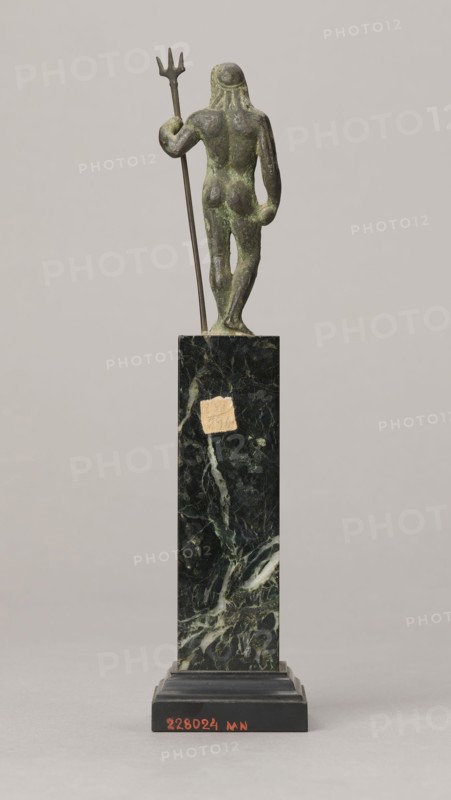 Figurine of Poseidon, unknown Roman workshop. Figurine of Poseidon standing in contrapposto, resting on a trident. The weight of the body rests on the left leg; the right is slightly moved back, the foot bent at the toes. The body is nude, correctly modeled, with clearly defined abdominal muscles. In the left hand the figure holds a trident; the right hand is lowered at the side of the body, holding a cylindrical object at hip level. The head is slightly bowed. The face is framed by long hair and a beard. 1st century, Roman Empire 27 BC–476. Bronze figurine; height 8.7 cm, width 3.5 cm, depth 1.8 cm, weight 0.362 kg with base., A small bronze figurine with a greenish patina stands on a tall dark green marble plinth; the figure is shown from the back holding a long thin trident in one hand, with a rounded head and stylized hair, and a weathered surface; the plinth has lighter veining and a small patch of lighter material, and rests on a stepped black base; the background is a pale neutr...