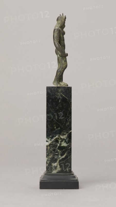 Figurine of Poseidon, unknown Roman workshop. Figurine of Poseidon standing in contrapposto, resting on a trident. The weight of the body rests on the left leg; the right is slightly moved back, the foot bent at the toes. The body is nude, correctly modeled, with clearly defined abdominal muscles. In the left hand the figure holds a trident; the right hand is lowered at the side of the body, holding a cylindrical object at hip level. The head is slightly bowed. The face is framed by long hair and a beard. 1st century, Roman Empire 27 BC–476. Terracotta figurine, height 8.7 cm, width 3.5 cm, depth 1.8 cm, weight 0.362 kg with base., A small greenish bronze figure stands in profile atop a tall dark green and black marble plinth with lighter green and white veining, the figure showing a textured mane of hair and a slender body with one arm bent and the other hanging, the plinth resting on a stepped black base against a light gray background.