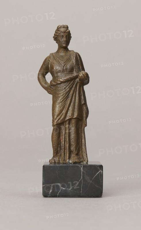 Venus figurine, unknown Roman workshop, Roman period, Roman Empire 27 BC–476, figurine, height 7.4 cm, width 2.8 cm, depth 2.5 cm, A small bronze-colored statue of a standing female figure wearing a draped garment, shown frontally on a dark gray marble base with white veins; the figure has one arm bent with a hand on her hip and the other arm holding a round object near her waist, with visible textured surfaces and folds in the clothing, and the background is a smooth light beige-gray.