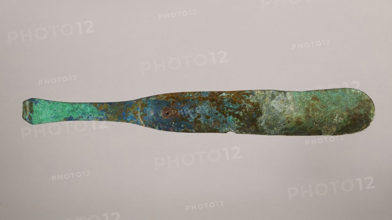 Scraper, metal scraper with a typical slightly curved shape consisting of a handle and a blade with a slightly rounded end. 6th–4th century BC, Rome, tool. Height 4.5 cm, width 28 cm, depth 11 cm, weight 0.11 kg., A long, thin metallic object with an irregular, flattened paddle shape lying horizontally against a plain light gray background; the surface shows mottled, weathered patina in shades of green, turquoise, blue, brown, and hints of rust, with textured patches and spots, a slightly tapered handle on one end and a broader rounded end on the other, and small areas of roughness and uneven edges along its length.
