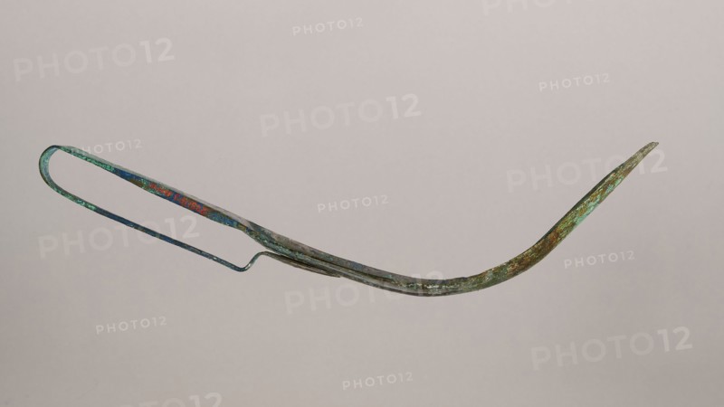 Scraper, metal scraper with a typical, slightly curved shape consisting of a handle and a blade with a slightly rounded end. 6th–4th century BC, possibly Rome; tool. Height 4.5 cm, width 28 cm, depth 11 cm, weight 0.11 kg., A slender curved metal object with a looped handle at one end and a tapered pointed tip at the other, showing surface irregularities and corrosion. The metal displays colors including green, turquoise, blue, brown, and hints of rusty orange, with mottled patches and areas of patina. The object has a narrow elongated body with a slight S-shaped bend near the handle and a thin supporting arm running along its length. The background is a plain light gray-beige.