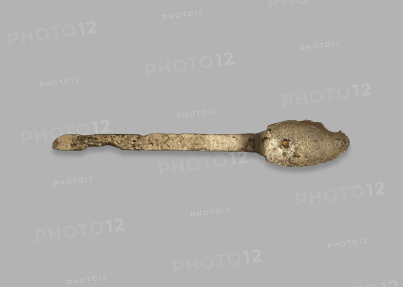 Medical tool fragment of strigil?, tool, height 12.8 cm, A single worn metal spoon lies horizontally against a plain light gray background; the spoon has a long, irregularly textured handle and an oval bowl with rough, pitted surfaces and an uneven edge, showing a tarnished metallic appearance in shades of dull gold, bronze, brown, and darker grayish spots.