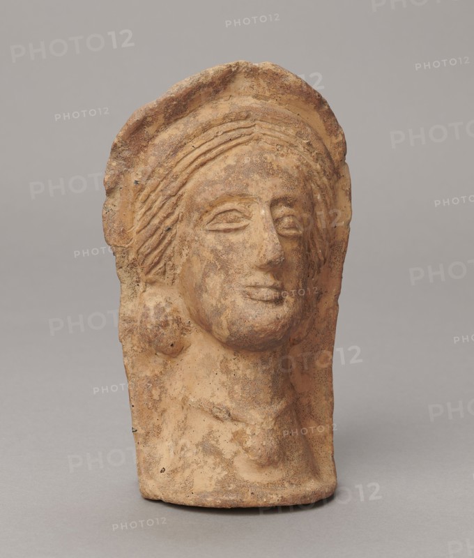 Votive head, 3rd–2nd century BC, Central Italy; sculpture; height 14.5 cm, width 9 cm, depth 8.5 cm, weight 0.5 kg; authenticity uncertain., A weathered terracotta sculpture head with a rounded top and stylized hair, displayed against a neutral gray background; the surface shows a rough texture with visible erosion and small pits, and the colors visible include shades of tan, light brown, reddish-brown, and darker brown speckling; the facial features are simplified with almond-shaped eyes, a pronounced nose, and full lips, and there is a suggestion of hair or a headdress framing the face and falling to the sides, with a short neck and a flattened back.
