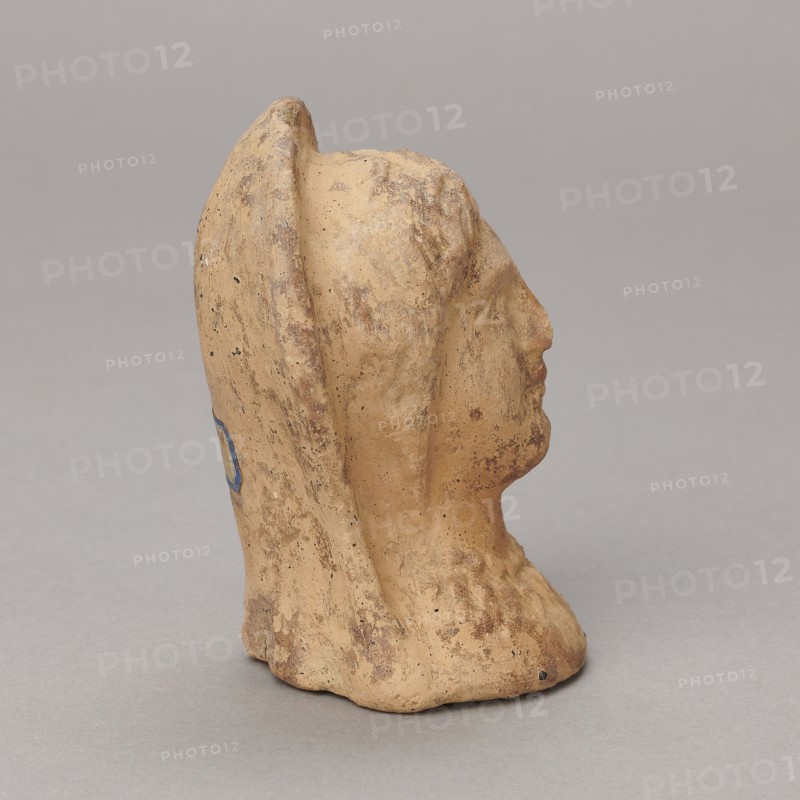 Ex voto, Madeyski, Antoni 1862–1939 - 30 BC; Hellenistic period; Southern Etruria, Latium ; bas-relief; height 9.5 cm, width 7 cm, depth 6.3 cm; weight 0.16 kg, A sculpted head in a beige to light tan clay color is shown in profile against a neutral gray background, with visible rough texture and patches of darker brown discoloration, and a small area of blue on the rear side; the face has a defined nose and chin, the head is draped with a raised hood or veil, and the base appears irregular and worn.