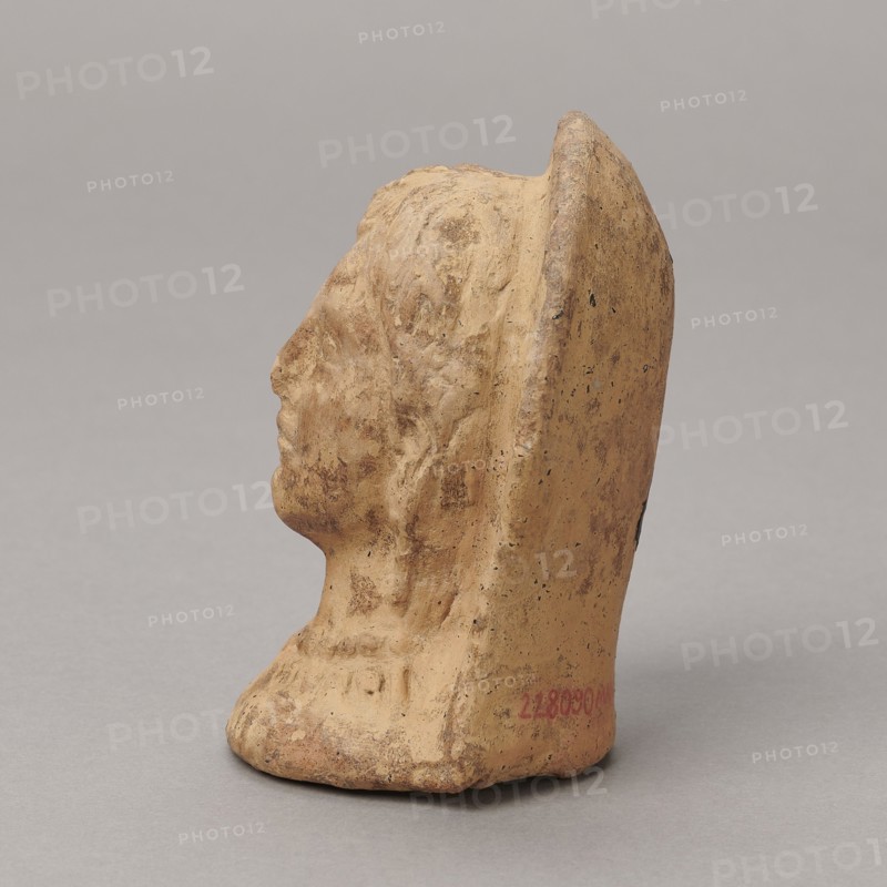 Ex voto, bas-relief, Hellenistic period, 332–30 BC; Southern Etruria, Latium ; height 9.5 cm, width 7 cm, depth 6.3 cm, weight 0.16 kg., A small sculpted profile of a human head and neck carved from a beige to light tan material with areas of darker brown patina and speckling, seen from the left side against a neutral gray background; the figure wears a rounded headdress or back piece that rises behind the head, and the surface shows textured tool marks and weathered roughness with subtle shadows and highlights.