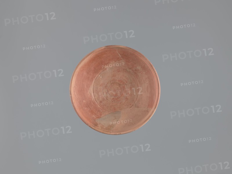 Plate, unknown Aswan workshop; flat plate on a low, ring-shaped foot. Rim poorly defined, rounded at the top with a vertical, slightly concave outer wall. Surfaces covered with red engobe and polished. Decoration executed by engraving and imprinting: three concentric grooves on the inner bottom, a roulette motif between the two outer grooves, and a central stamped cross in the form of a crux gemmata. Made of pink kaolin clay. 6th–7th century. Find location: Edfu. Vessel plate. Height 5.0–5.3 cm; diameter 26.8 cm rim, 13.7 cm bottom. Materials/technique: plates, Aswan pottery, Egyptian pottery, Egyptian sigillata, tableware, Coptic vessels, stamp impressions, stamped decoration, roulette decoration, kaolin clay., A round shallow dish with a worn terracotta surface sits against a smooth muted gray background; the dish shows circular ridges and areas of surface wear with a few lighter patches and subtle variations in reddish-brown and pale beige tones.