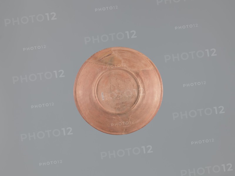 Plate, unknown Aswan workshop; flat plate on a low ring-shaped foot. Rim poorly defined, rounded at top, with a vertical, slightly concave outer wall. Surfaces covered with red engobe and polished; made of pink kaolin clay. Decoration by engraving and imprinting: three concentric grooves on inner bottom, roulette motif between outer grooves, central stamped cross in form of a crux gemmata. Egyptian pottery Egyptian sigillata, tableware/Coptic vessel; stamp impressions, stamped and roulette decoration. 6th–7th century; findspot Edfu. Vessel/plate; height 5.0–5.3 cm; diameter rim 26.8 cm; diameter bottom 13.7 cm., A round, shallow dish with a worn, matte surface in shades of terracotta and muted orange sits centered against a flat, uniform background of cool gray. The dish shows subtle variations in color with areas of lighter peach and darker rust, and faint circular ridges and scuff marks across its face. There are a few triangular repair lines and slightly darker patches near the rim,...