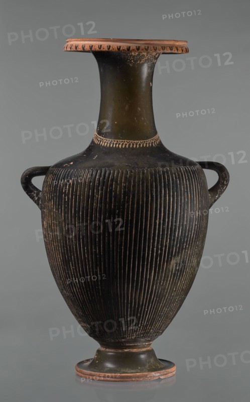 Black-finned hydria with fluting, unknown Apulian workshop. Hydria with fluting belly. Red clay. Black and shiny engobe firnis. The rim widens and turns outward, decorated with a painted ornament called an ovoid oxeye. The neck is long, cylindrical, decorated with a gilded garland painted at the base. The shoulders are clearly defined. The belly is oval, tapering towards the base, covered with fluting, impressed from the mold. The handles are cylindrical, attached vertically at shoulder height. Wide base. Preservation condition: cracked engobe in places, gilding worn away. 320–310 BC, Apulia, hydria vessel. Height 57 cm, width 24.7 cm., A tall ceramic vase with a narrow flared neck and a wide rounded body, featuring a pair of horizontal loop handles near the shoulder; the vase surface shows a ribbed texture on the body and a smoother neck and base with small areas of wear and speckling. The colors visible include deep black on most of the body and neck, earthy terracotta on the rim and...