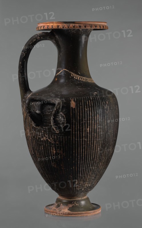 Black-finned hydria with fluting, unknown Apulian workshop. Hydria with fluting belly. Red clay. Black and shiny engobe firnis. The rim widens and turns outward, decorated with a painted ornament called an ovoid oxeye. The neck is long, cylindrical, decorated with a gilded garland painted at the base. The shoulders are clearly defined. The belly is oval, tapering towards the base, covered with fluting, impressed from the mold. The handles are cylindrical, attached vertically at shoulder height. Wide base. Preservation condition: cracked engobe in places, gilding worn away. 320–310 BC, Apulia, hydria, vessel; height 57 cm, width 24.7 cm., A tall, narrow-handled ceramic vessel with a dark black surface and areas of worn terracotta color, featuring a rounded body that tapers to a small foot, a flared rim with a band of lighter terracotta around the edge, a curved single handle from rim to shoulder, a pair of small looped side handles on the shoulder, vertical incised lines covering the lo...