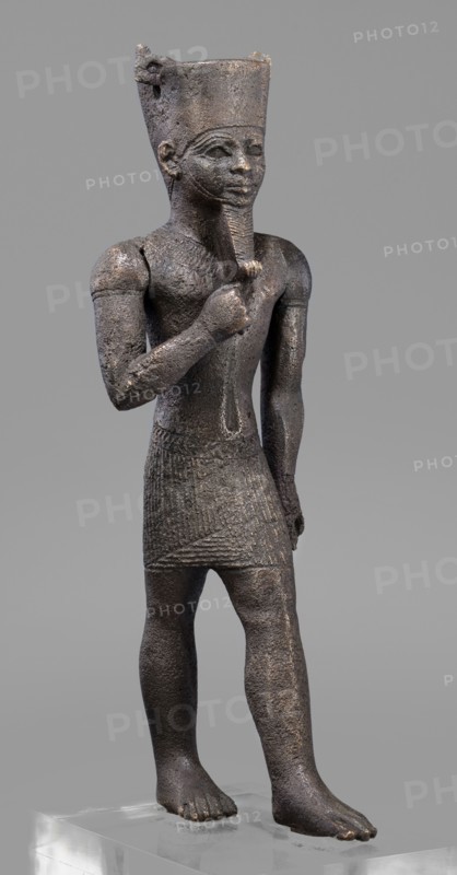Figurine of the god Amun, Amun mythology, Egyptian deities, male figurines, Egyptian art, Once holding a khepesh, ca. 1069–ca. 747 BC; Third Intermediate Period, Egypt, sculpture, height 19.6 cm, width 6.2 cm, A dark brown textured statue of a standing human figure on a clear rectangular base, wearing a tall flat-topped headdress and a short patterned skirt, with one arm bent so the hand touches a stylized beard and the other arm hanging by the side, the face showing almond-shaped eyes and a calm expression, the surface showing rough, worn areas and lighter brown highlights against a neutral gray background.