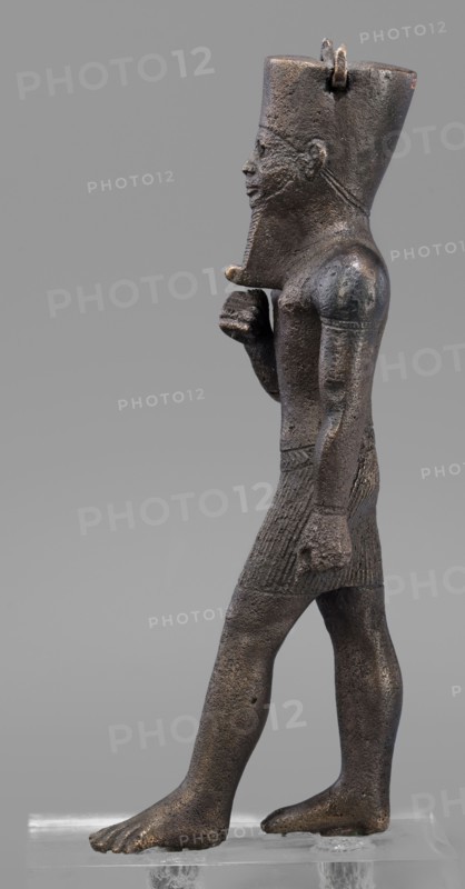 Figurine of the god Amun; maker, Amun mythology; Egyptian deity male figurine; once held a khepesh; ca. 1069–ca. 747 BC, Third Intermediate Period, Egypt; sculpture; height 19.6 cm, width 6.2 cm., A dark brownish bronze statue of a standing male figure shown in profile, facing left, wearing a tall headdress and a short textured skirt; the figure has a stylized beard and a pronounced jawline, a closed mouth, and a visible ear, with one arm bent and fist near the chest and the other arm hanging down with a clenched hand; the surface shows a rough, pitted texture with areas of lighter brown and subtle greenish or blackish patina; the figure stands barefoot on a clear rectangular support against a plain light gray background.