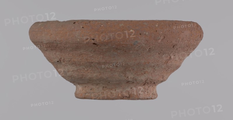 Bowl, Egyptian pottery, clay vessel, bowl, 332 BC–395 AD Ptolemaic–Roman Period, from Edfu; vessel; height 5 cm; rim diameter 11 cm; bottom diameter 5.7 cm., A worn, shallow bowl-like object with a rough, porous surface in shades of reddish-brown and muted orange, showing patches of darker brown and small areas of gray; the form has a flared rim, a gently curved body, and a narrower foot with uneven texture, with visible pits and small holes across its surface against a pale gray background.