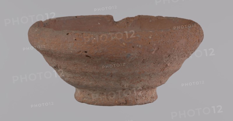 Bowl, unknown creator; Egyptian pottery vessel made of clay; bowl; Ptolemaic Period–Roman Period 332 BC–395 AD; from Edfu; height 5 cm; rim diameter 11 cm; bottom diameter 5.7 cm., A weathered clay bowl with a rough, pitted surface sits against a plain light gray background; the bowl is mostly reddish-brown with patches of darker brown and small flecks of lighter beige, showing irregular texture and small holes, a slightly flared rim with uneven edges, a rounded body that tapers to a short, slightly raised foot, and visible surface variations and chips.