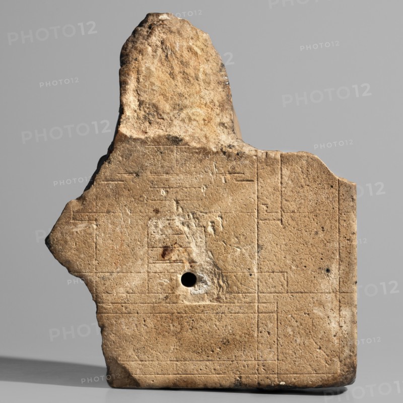 Sculptural model — head of Nectanebo I or II. Sculpture model of a ruler's head in full relief attached to a flat slab background fragmentarily preserved on the right. The ruler wears a nemes covering half the forehead, decorated at the front with a uraeus whose body is curled in a horizontal zigzag similar to a figure-eight with a straight tail. The nemes bears twisted ram's horns. The face is oval and plump with rounded chin and cheeks; small mouth with upper and lower lips of equal width and slightly upturned corners; long nose; small, close-set, slightly slanting eyes with upper and lower eyelids highlighted by thin lines; wide eyebrows. Very careful modeling with precisely executed details; auxiliary lines visible on the back. With base. Height 13 height 11 cm, width 9 cm, depth 5 cm, weight 0.338 kg. 380–343 BC; Late Period, 30th Dynasty reign of Nectanebo I or II; Egyptian sculpture., A weathered stone fragment with an irregular top edge and a roughly triangular protrusion on on...