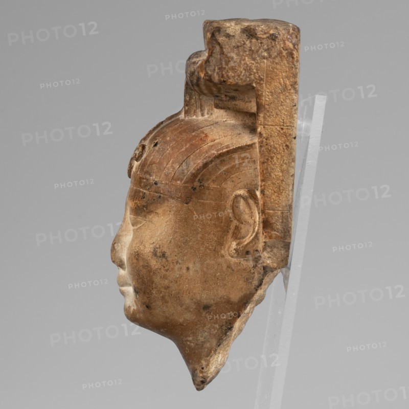 Sculptural model - head of Nectanebo I or II. Sculpture model in stone, atef crown iconographic, uraeus iconographic, nemes scarf iconographic, horns iconographic. The ruler's head is in full sculpture connected to a flat slab background fragmentarily preserved, only on the right side. The ruler wears a nemes covering half his forehead; its front is decorated with a uraeus whose body is curled in a horizontal zigzag similar to a figure-eight, tail straight. The nemes bears twisted ram's horns. Face oval and plump with round chin and cheeks; small mouth with upper and lower lips of equal width and slightly upturned corners; long nose; small, close-set, slightly slanting eyes with upper and lower eyelids highlighted by thin lines; wide eyebrows. Very careful modeling with precisely executed details. Auxiliary lines visible on the back. With base. Height 13 cm object dimensions: height 11 cm, width 9 cm, depth 5 cm, weight 0.338 kg. Date: 380–343 BC; Late Period, 30th Dynasty reign of Nec...
