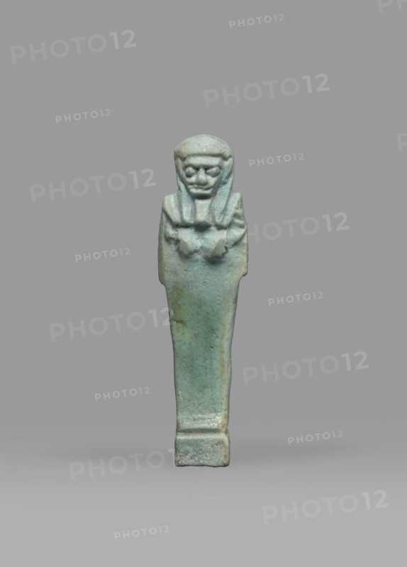 Ushabti, Mummy-shaped funerary figurine shabti, standing on a base, back supported by a pillar/pilaster. Arms crossed on the chest, right folded over left; both hands holding hoes. A bag is on the back, slung over the left shoulder on a single string. Wearing a three-part smooth wig with the back strand longer than the front strands; long, straight, wide beard. Large eyes, wide nose, mouth set in a grimace, very large ears. No inscription. Late Period, Egypt, 664–332 BC. Height 7.7 cm; width 2.1 cm., A small carved figurine stands upright against a plain gray background, showing a stylized human form with a headpiece and a long, tapered body resting on a short rectangular base; the surface is predominantly a bluish-green with areas of muted green and pale gray, and the figure's facial features and folded arms are roughly modeled with subtle texture and weathered patina.