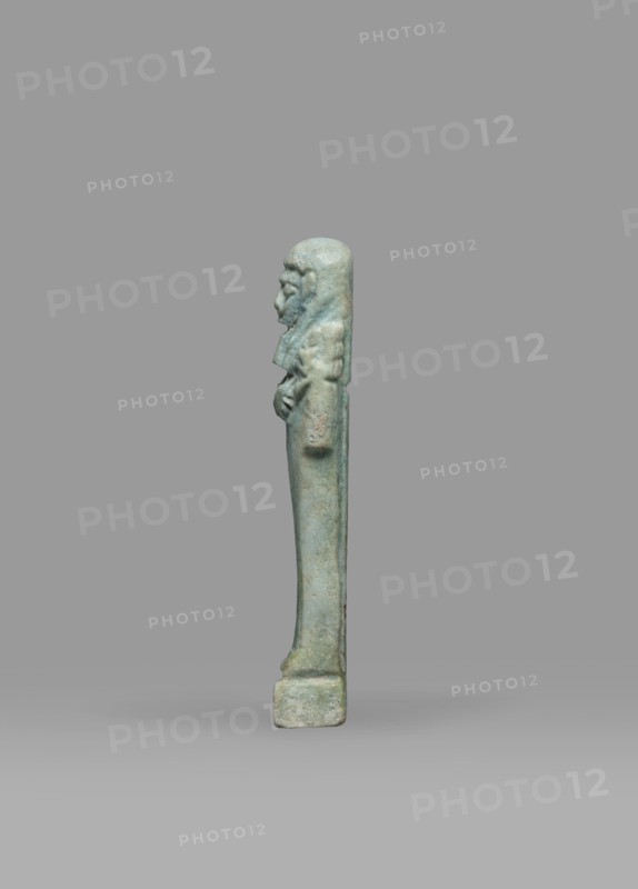 Ushabti, Mummy-shaped funerary figurine shabti standing on a base, back supported by a pillar/pilaster. Arms crossed on the chest, right folded over left; both hands holding hoes. A bag is on the back, slung over the left shoulder on a single string. Wearing a three-part smooth wig, the back strand longer than the front strands. Long, straight, wide beard; large eyes, wide nose, mouth set in a grimace, very large ears. No inscription. Late Period, Egypt, 664–332 BC. Height 7.7 cm, width 2.1 cm., A tall, narrow statuette shown in profile against a plain background, rendered in greenish-blue tones with areas of paler and darker green; the figure stands on a small rectangular base and has a long, slender body with a straight posture; the head is rounded with visible facial features in low relief and a headdress or hair that falls down the back; the arms are bent and held close to the torso with hands resting near the abdomen; the surface shows a weathered, slightly rough texture with subt...