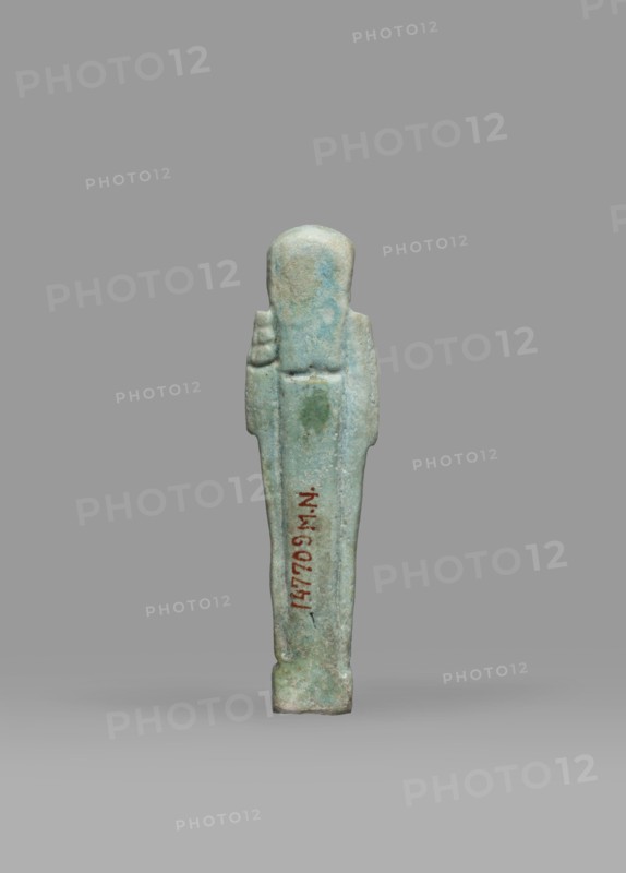 Ushabti, Mummy-shaped funerary figurine shabti, standing on a base, back supported by a pillar/pilaster. Arms crossed on the chest, the right folded over the left; both hands holding hoes. A bag is on the back, slung over the left shoulder on a single string. Wears a three-part smooth wig back strand longer than front strands. Long, straight, wide beard. Large eyes, wide nose, mouth set in a grimace, very large ears. No inscription. Late Period, Egypt, 664–332 BC. Height 7.7 cm, width 2.1 cm., A small upright figurine with a smooth rounded head and simplified body stands against a plain gray background; the figure is predominantly light blue-green with areas of pale turquoise and subtle greenish patches, and there is a narrow darker reddish-brown vertical detail on its front; the surface appears worn and slightly pitted, and the base is a muted gray-green.