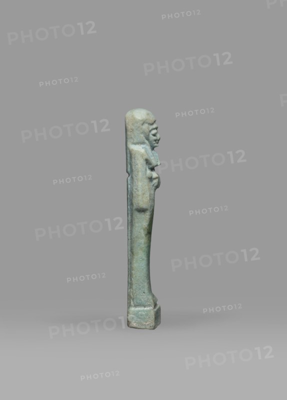 Ushabti, Mummy-shaped funerary figurine shabti, standing on a base, back supported by a pillar/pilaster. Arms crossed on the chest, the right folded over the left; both hands holding hoes. A bag is on the back, slung over the left shoulder on a single string. Wearing a three-part smooth wig with the back strand longer than the front strands; long, straight, wide beard. Large eyes, wide nose, mouth set in a grimace, very large ears. No inscription. Late Period, 664–332 BC, Egypt. Height 7.7 cm; width 2.1 cm., A small upright statuette of a human figure shown in profile against a plain gray background; the figure is narrow and elongated with a rounded head, pronounced facial features, and arms held close to the torso, standing on a rectangular base. The surface shows a weathered, mottled greenish-blue color with areas of lighter and darker green and hints of brownish-gray patina. The background is a uniform medium gray with a slightly lighter gray ground beneath the base.