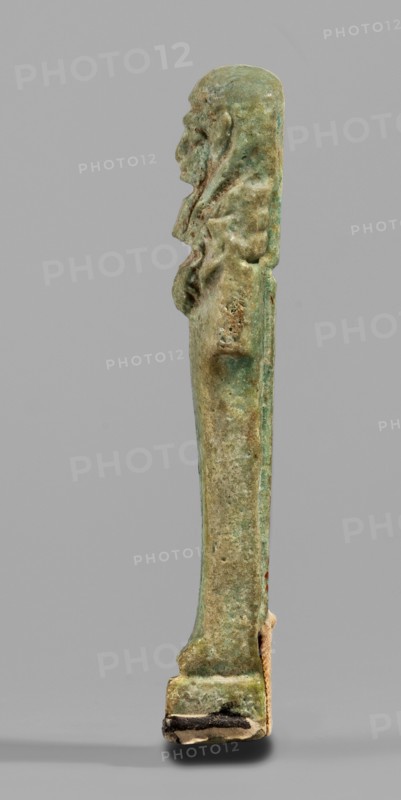 Ushabti, Mummy-shaped funerary figurine, standing on a base with back supported by a pillar/pilaster. Arms crossed on the chest, right folded over left; both hands holding hoes. A bag is slung over the left shoulder on a single string on the back. Wears a three-part smooth wig back strand longer than front strands; long, straight, wide beard. Large eyes, wide nose, mouth set in a grimace, very large ears. No inscription. Late Period, 664–332 BC, Egypt. Shabti; height 7.7 cm, width 2.1 cm., A slender vertical statuette stands against a plain light gray background, showing a side profile of a human figure with a pronounced head, facial features, and hair or headdress that extends down the back; the surface appears weathered and textured with irregularities and small holes. The object displays a mix of greenish and bluish patina with areas of brown and tan discoloration, darker green and brown spots, and lighter beige or off-white patches near its base. The lower end rests on a small rect...