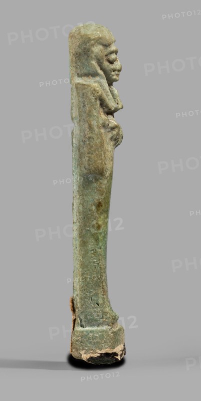Ushabti, Mummy-shaped funerary figurine standing on a base, back supported by a pillar/pilaster. Arms crossed on the chest, right folded over left; both hands holding hoes. A bag is slung over the left shoulder on a single string on the back. Head wears a three-part smooth wig with the back strand longer than the front strands. Long, straight, wide beard. Large eyes, wide nose, mouth set in a grimace, very large ears. No inscription. Late Period, 664–332 BC, Egypt. Shabti. Height 7.7 cm, width 2.1 cm., A vertical, elongated statuette in shades of green and brown with a weathered, mottled surface, showing a stylized human figure in profile near the top with a pronounced nose and chin, arms folded against the chest, and a rounded head; the base is wider, rough and chipped with visible beige and dark brown areas, and the background is a flat neutral gray.