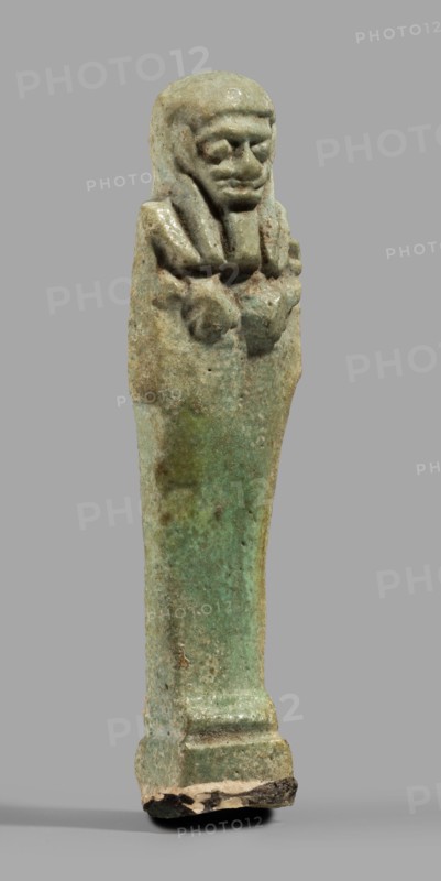 Ushabti, Mummy-shaped funerary figurine standing on a base, back supported by a pillar/pilaster. Arms crossed on the chest, the right folded over the left; both hands holding hoes. A bag is on the back, slung over the left shoulder on a single string. Wearing a three-part smooth wig with the back strand longer than the front strands. Long, straight, wide beard; large eyes, wide nose, mouth set in a grimace, very large ears. No inscription. Late Period, Egypt, 664–332 BC. Shabti; height 7.7 cm, width 2.1 cm., A small statuette of a standing figure with a long body and a stylized head and shoulders, shown against a plain gray background; the figure appears to wear a headdress with a fringe and has a defined face with a pronounced chin and brow, and arms crossed over the chest; the surface shows a weathered, mottled texture with visible pitting and flaking; colors visible include shades of green, turquoise, pale blue, beige, brown, and touches of darker brown or black at the base; the bas...
