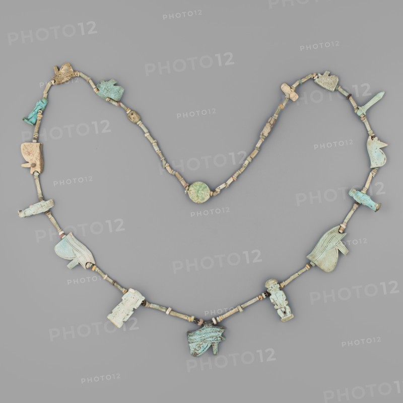 Necklace with amulets, udjat eyes, tit knots, Ptah the dwarf, Toeris, necklaces, columns, auletes separated by tubular and disc-shaped beads: 8 udjat eyes, Isis knot, Toeris, papyrus column, Ptah the dwarf, 2 anthropomorphic deities, 2 plaques; ca. 1550–332 BC, New Kingdom–Late Period, Egypt, amulet, height 70 cm, A beaded necklace arranged on a gray background showing a string of small cylindrical and disc-shaped beads in beige, tan, and light brown, interspersed with a few larger carved pendants in shades of pale blue, turquoise, greenish-blue, cream, and light beige; the pendants include a few stylized animal and figure shapes and some abstract carved forms, each pendant displaying weathered surfaces and subtle color variations; the overall palette includes gray from the background and the beads’ beige and tan tones alongside the blue and green hues of the larger pieces.