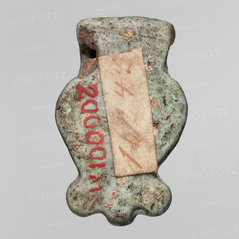 Amulet - head of the goddess Hathor;, Hathor myth.; Uraeuses; Face of the goddess Hathor in a Hathor wig with a wreath of Uraeuses; ca. 1069–ca. 656 BC, Third Intermediate Period, Egypt; amulet; height 2 cm, width 1.6 cm, An irregularly shaped small object with a weathered, pitted surface appears centered against a plain light gray background; the object shows a predominantly pale greenish-blue color with areas of brownish discoloration and darker green speckling, a rectangular beige paper strip adhered across its middle, and a few bright red painted marks along one side; the shape includes a narrower top, a rounded body with indentations at the sides, and a scalloped lower edge.