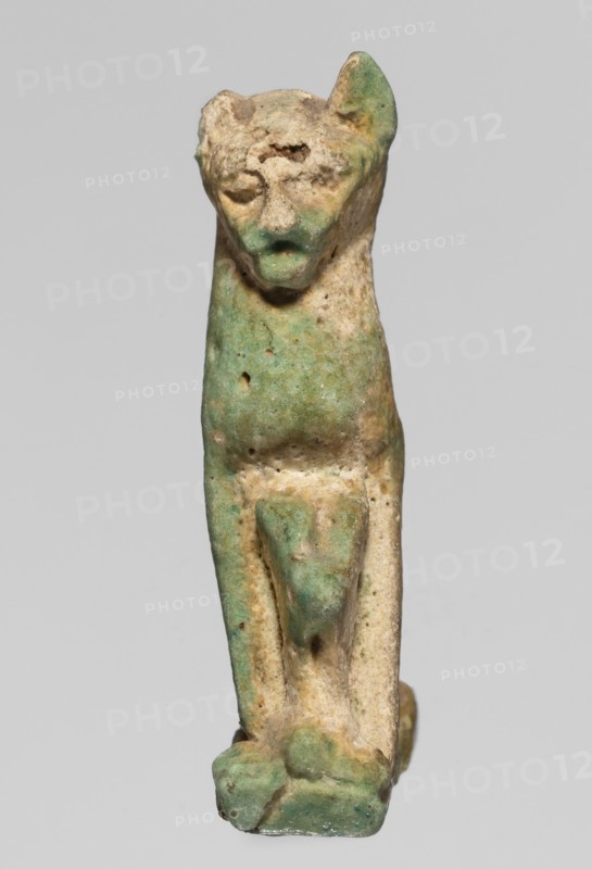 Amulet — cat; maker, cats; pendant; amulet; material unspecified; ca. 945–525 BC 22nd–26th Dynasty, Egypt; height 3.3 cm, width 2 cm., A small weathered figurine of an animal-like form with a rounded head, pointed ears, and a simplified face, standing upright on a narrow rectangular base; the surface shows a mottled mix of green and beige tones with areas of darker and lighter green patina, pitted and rough textures, and some smoother worn spots, with the limbs and body rendered in a stylized, blocky manner and the overall appearance suggesting age and abrasion.