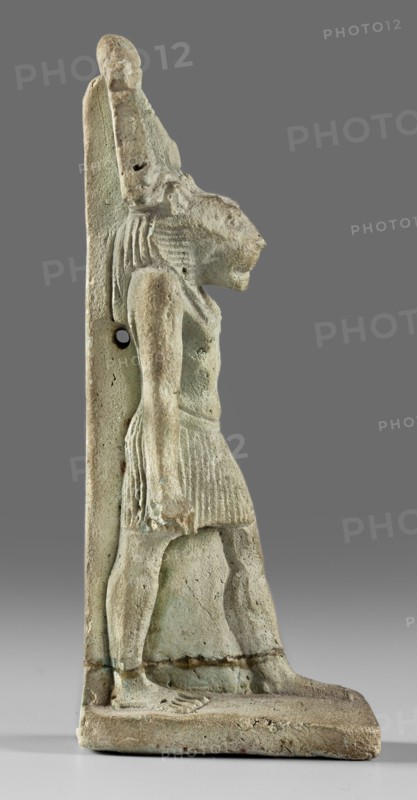 Amulet - god Mahes; figurine of a striding lion-headed male deity wearing an atef crown, dressed in a pleated loincloth; pillars on the back; amulet; Mahes mythology; 1st century, Roman period, Egypt; height 7.4 cm, width 2.2 cm., A weathered carved figure in profile standing on a rectangular base, showing a humanoid body with a feline head and an upright back pillar; the figure wears a pleated skirt and has a defined arm hanging alongside the torso, with visible grooves suggesting hair or a headdress; surface texture is pitted and rough with areas of erosion and small holes; colors include pale greenish-blue, beige, light gray, and muted brown tones, with darker gray used for the background.