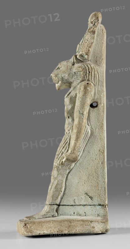 Amulet - god Mahes. Figurine of a striding lion-headed male deity wearing an atef crown, dressed in a pleated loincloth; pillars on the back. Egyptian deity Mahes mythology. Roman period, 1st century CE, Egypt. Material: amulet. Height 7.4 cm; width 2.2 cm., A side view of a vertical carved figure in a muted beige and pale green material, showing a human-like body with a feline head and a tall headdress, a few areas of darker patina and weathering, a group of carved ridges suggesting hair or mane, arms held down along the sides, a tapered garment with vertical grooves, a flat rectangular base supporting the figure, and a plain gray background.
