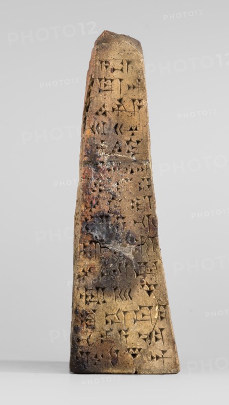 Votive obelisk with an inscription of King Ashurbanipal forgery with a copy of an ancient text, cuneiform writing, Ashurbanipal king of Assyria; ca. 668–ca. 631 BC, votive offering obelisk. Clay obelisk tapering toward the top with cuneiform text inscribed on all four faces. Dating of copied inscriptions: c. 668–c. 631 BC Neo-Assyrian period; object itself dated to 2nd half 19th century–1st half 20th century forgery. Inscription based on texts from Iraq Quyunjik, Nineveh. Function: dedication/document. Dimensions: height 13.1 cm; width 3.7 cm; thickness 4.2 cm., A tapered, upright stone object with a rough, weathered surface stands against a neutral light gray background; the object is primarily tan and brown with patches of darker brown and black discoloration and small areas of reddish coloration, and its surface shows numerous small triangular and wedge-shaped indentations arranged across the face; the top is irregular and slightly angled, the sides slope inward toward the top, and ...
