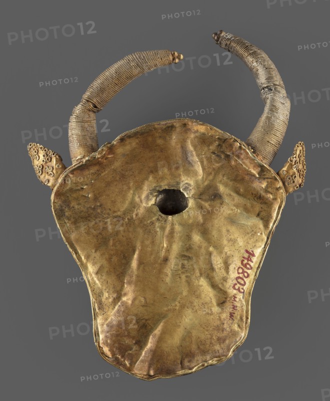 Protoma; bull's head relief mask in gold, made using repoussé with granulation and filigree. Central part with nostrils features granulation and filigree; cheeks and top of head feature filigree only. Eyes, nostrils, and muzzle are repoussé and bordered with filigree. Small ears and horns are plastically modeled horns soldered on. Reverse closed with an undecorated gold sheet with an irregular hole in about 2/3 of its height from removal of a fastening element. Materials/technique: goldsmithing. Culture/style: Elamite Iranian art. Date: ca. 1450–ca. 1100 BC, Middle Elamite period, — alternate datings include Achaemenid period 600–539 BC and arguments for second half of the 2nd millennium BC. Place of origin: Elam/Iran . Function: protoma decorative/attachment. Dimensions: height 9.5 cm, width 7.8 cm, depth 2.5 cm; weight 48 g., An object with a large irregularly shaped metallic surface that appears crumpled and worn, showing a tarnished golden color with areas of darker brown and subtl...