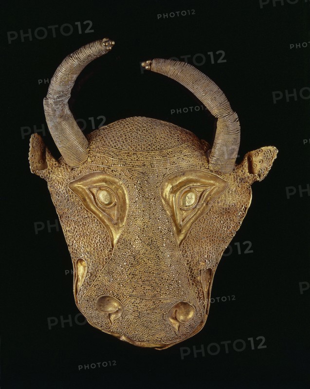 Protoma bull head relief mask. Gold; repoussé, granulation, filigree, soldering. Central nostril area with granulation and filigree; cheeks and top with filigree; eyes, nostrils and muzzle repoussé and bordered with filigree. Small ears and plastically modeled horns horns soldered on. Reverse closed with an undecorated gold sheet with an irregular hole in two-thirds of its height from removal of a fastening element. Ca. 1450–1100 BC Middle Elamite period ?; alternative proposed datings include Achaemenid period 600–539 BC and second half of the 2nd millennium BC. Origin: Elam/Iran. Decoration. Height 9.5 cm; width 7.8 cm; depth 2.5 cm; weight 48 g., A golden decorative mask in the shape of an animal head set against a black background, featuring a textured surface made of many small circular patterns and smooth raised areas, with a pair of curved horns wrapped in a fine coil-like texture, prominent sculpted eye shapes and a pronounced snout, and visible colors including gold tones and ...