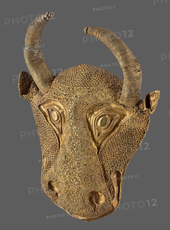 Protoma; bull's head relief mask made in gold using repoussé, filigree, and granulation. Central nostril area features granulation and filigree; cheeks and top of head use filigree. Eyes, nostrils, and muzzle are repoussé and bordered with filigree. Small ears and horns are plastically modeled horns soldered on. Reverse closed with an undecorated gold sheet with an irregular hole in two-thirds of its height from removal of a fastening element. Ca. 1450–1100 BC Middle Elamite period, — alternative dating to Achaemenid period 600–539 BC has been proposed; arguments also presented for second half of 2nd millennium BC. Elam, Iran. Decoration. Height 9.5 cm; width 7.8 cm; depth 2.5 cm; weight 48 g., A gold-colored decorative mask shaped like an animal head with two curved horns, a textured surface made of many small circular elements, prominent eye areas with raised almond-shaped surrounds, a long tapered snout with rounded nostril shapes, and small ear-like projections on either side, show...