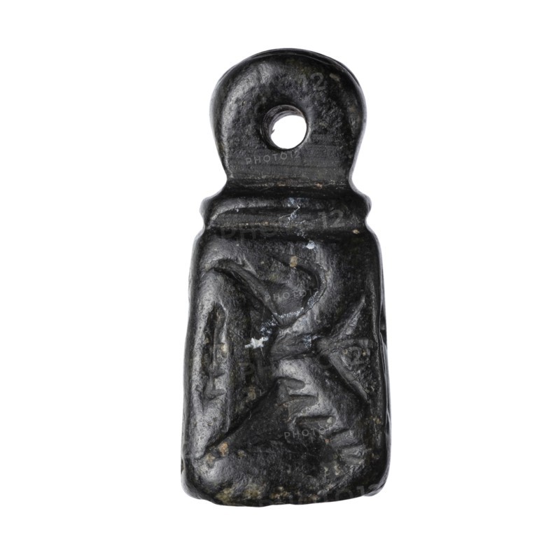 Stamp seal with figural representations. Stamp seal in the form of a truncated pyramid on a square base, terminated with a round handle. The four side walls and the base are decorated with engraved, stylized figurative representations: two opposite sides depict a rider on a horse; one of the other sides depicts a striding figure holding a staff topped with a star; the opposite wall depicts an animal being attacked by a bird of prey. A winged animal is engraved on the seal's base. Glyptics; figural motifs; Eastern Gallic scenes. 9th–6th century BC, Iron Period. Northwestern Syria; Near East. Seal. Height 2.8 cm; width 1.3 cm; depth 1.4 cm. Seal impression carrier length 8.5 cm; width 4.4 cm., A small dark pendant shaped like a flattened rectangle with a rounded top and a circular hole near the top, showing a textured surface with carved or worn shallow grooves and irregularities; the visible colors are predominantly black with a few spots of gray and small flecks of brown or beige.