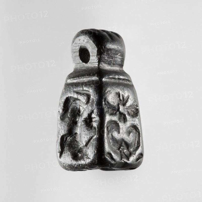 Stamp seal with figural representations. Glyptic, stamp seal in the form of a truncated pyramid on a square base, terminated with a round handle. The four side walls and the base are engraved with stylized figurative representations: two opposite sides show a rider on a horse; one of the other sides depicts a striding figure holding a staff topped with a star; the opposite wall depicts an animal being attacked by a bird of prey. A winged animal is engraved on the seal's base. Iron Age, 9th–6th century BC; Northwestern Syria, Near East. Seal impression carrier dimensions: length 8.5 cm, width 4.4 cm. Seal dimensions: height 2.8 cm, width 1.3 cm, depth 1.4 cm., A small dark gray and black carved object with a rounded top that has a hole through it, a vertical ridge down the center, and a textured lower portion showing carved floral and curved decorative motifs on either side, set against a light gray background.