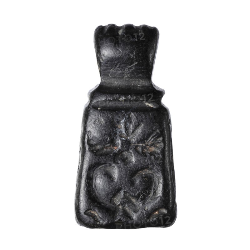 Stamp seal with figural representations. Glyptic stamp seal in the form of a truncated pyramid on a square base, terminated with a round handle. The four side walls and the base are decorated with engraved, stylized figurative representations: two opposite sides depict a rider on a horse; one side depicts a striding figure holding a staff topped with a star; the opposite wall depicts an animal being attacked by a bird of prey. A winged animal is engraved on the seal's base. Iron Age, 9th–6th century BC. Northwestern Syria; Near East. Material: seal. Dimensions: height 2.8 cm, width 1.3 cm, depth 1.4 cm; impression carrier dimensions: length 8.5 cm, width 4.4 cm., A dark, irregularly shaped carved object with a glossy surface, showing a stepped top section and a wider lower body; the surface has carved, rounded motifs including a heart-like form and a cross-like shape, with visible tool marks and small areas of lighter wear; predominant colors are black with touches of gray and faint br...