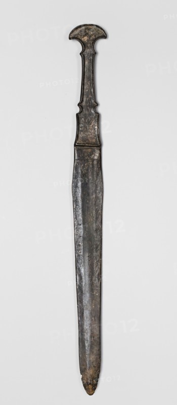 Dagger, unknown Luristan workshop. Dagger with a cast blade and a mushroom-shaped hilt with a rimmed mount; the raised edges of the mount served to hold a lost hilt casing, possibly of bone, wood, horn, or stone. The hilt shaft is richly profiled and flows directly into the blade with a centric point. Behind the short, rectangular base, blunted on both sides, the double-edged blade tapers evenly toward the tip. A slightly convex ridge runs through the center of the blade and splits along one-third of its length toward the base, creating a triangular flat field. 9th–6th century BC; Iron Period II 1st half of the 1st millennium BC. Luristan, Iran. Weapon. Height 33.8 cm; width 2.9 cm; depth 0.8 cm; weight 194 g., A long vertical metallic object with an elongated straight blade and a distinct handle at the top, displayed against a light gray background. The handle has a rounded top and a narrow neck with sculpted inward curves, showing an aged, slightly irregular surface. The blade is str...