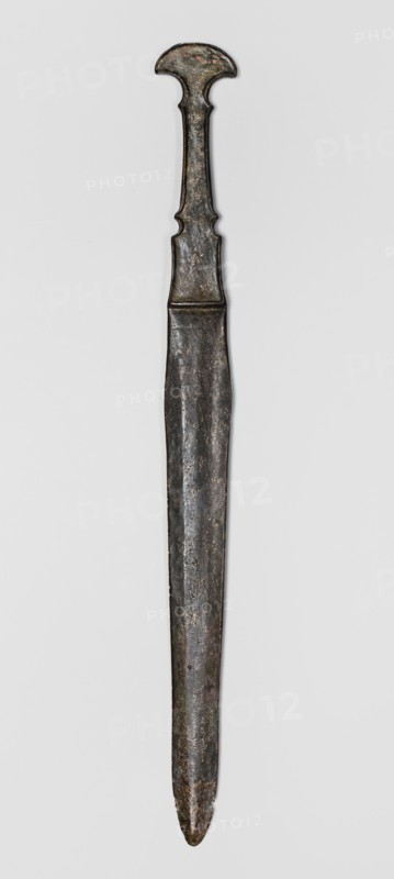 Dagger, unknown Luristan workshop. Dagger with a cast blade and a mushroom-shaped hilt with a rimmed mount; the raised edges of the mount served to hold a lost hilt casing possibly bone, wood, horn, or stone. The hilt shaft is richly profiled and flows directly into the blade with a centric point. Behind the short, rectangular base, blunted on both sides, the double-edged blade tapers evenly towards the tip. A slightly convex ridge runs through the center of the blade, which splits along one-third of its length towards the base, creating a triangular flat field. 9th–6th century BC; Iron Period II 1st half of the 1st millennium BC. Luristan, Iran. Weapon. Height 33.8 cm; width 2.9 cm; depth 0.8 cm; weight 194 g., A single elongated metal object with a narrow pointed end and a wider decorative top, shown against a light background; the object has a long straight shaft that tapers slightly toward the tip and displays an uneven, textured surface with signs of wear; the upper portion featur...